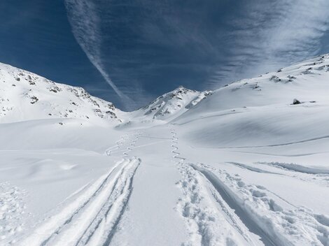 Eine verschneite Landschaft mit Spuren im Schnee und hohen Bergen. Der Himmel ist blau mit einigen Wolken.