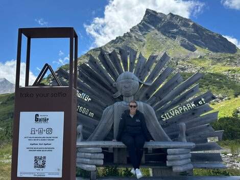 Eine Person sitzt auf einer großen Holzfigur mit Sonnenbrille. Im Hintergrund sind Berge und ein klarer blaue Himmel zu sehen.