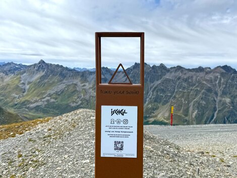 Ein Informationsschild auf einem Gebirgspfad mit Aussicht auf die umliegenden Berge. Der Himmel ist bewölkt und die Landschaft ist felsig.