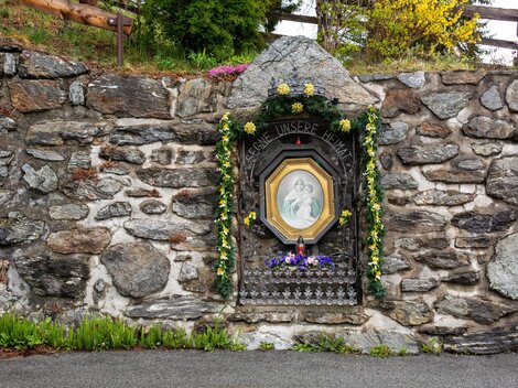 Eine Steinmauer mit einem dekorierten Bildnis in einer Nische. Das Bild ist mit Blumen geschmückt und zeigt eine religiöse Szene. | © Bernhard Gruber – Photography