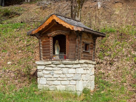 Eine kleine Holzkapelle steht auf einem Steinsockel in der Natur. Neben der Kapelle sind grüne Wiesen und Laub zu sehen. | © Bernhard Gruber – Photography