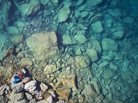Ein klarer, blauer See mit vielen großen Steinen am Grund. Eine Person liegt entspannt am Ufer.