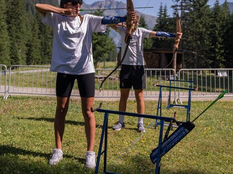 Two teenagers are practicing archery in a beautiful, natural setting. Mountains and trees are visible in the background.