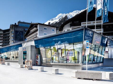 Ein modernes Gebäude mit großen Glasfenstern in einer Alpenlandschaft. Im Hintergrund sind schneebedeckte Berge und typische alpine Architektur zu sehen.