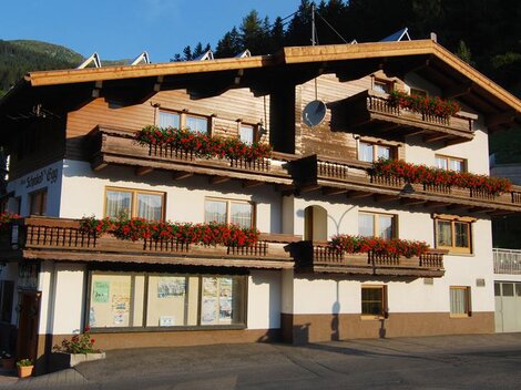 Ein traditionelles Holzgebäude mit vielen Balkonen, die mit roten Blumen geschmückt sind. Im Hintergrund sind grüne Berge zu sehen.