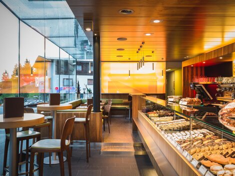 Ein modernes Café mit großer Fensterfront und einladender Atmosphäre. Die Theke ist mit verschiedenen Gebäcken und Getränken dekoriert.