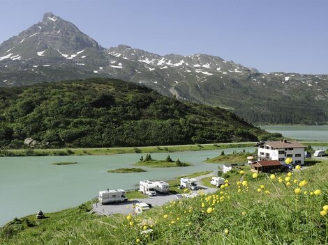 Eine malerische Landschaft mit einem ruhigen See und einem beeindruckenden Berg im Hintergrund. Am Ufer stehen Wohnmobile und ein Gasthaus umgeben von grünen Wiesen.