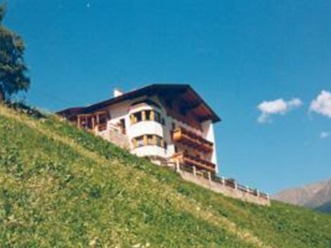 Ein schönes Haus auf einem Hügel, umgeben von grünen Wiesen. Der Himmel ist blau mit einigen Wolken.