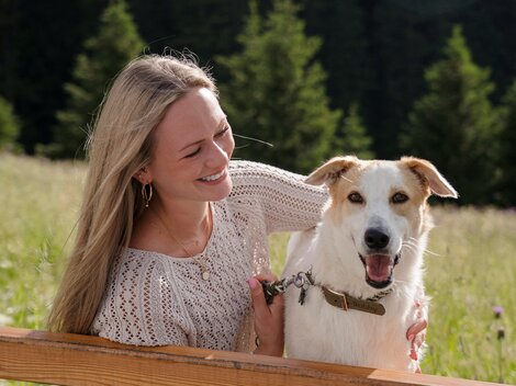 Eine Frau sitzt auf einer Bank und lächelt glücklich zu ihrem Hund. Der Hund schaut fröhlich zurück, umgeben von einer schönen, natürlichen Landschaft.