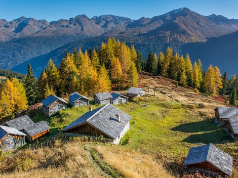 Eine malerische Berglandschaft mit mehreren Hütten und buntem Herbstwald. Im Hintergrund steigen majestätische Berge auf.