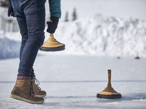 Eine Person steht auf einer gefrorenen Fläche und hält einen Holzblock in der Hand. Im Hintergrund sind schneebedeckte Hügel zu sehen.