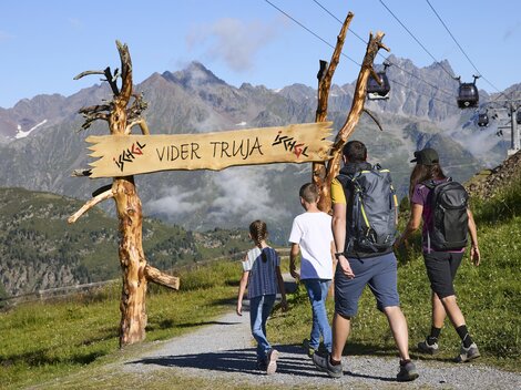 Eine Wandergruppe geht einen malerischen Weg zu einem Schild mit der Aufschrift „Vider Truja“. Im Hintergrund sind majestätische Berge und eine Seilbahn zu sehen.
