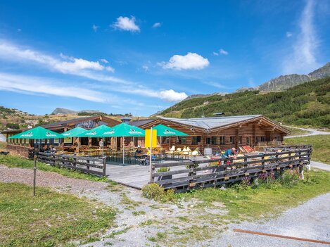 A cozy mountain cabin with a terrace and sun umbrellas. Surrounded by green meadows and mountains under a blue sky.