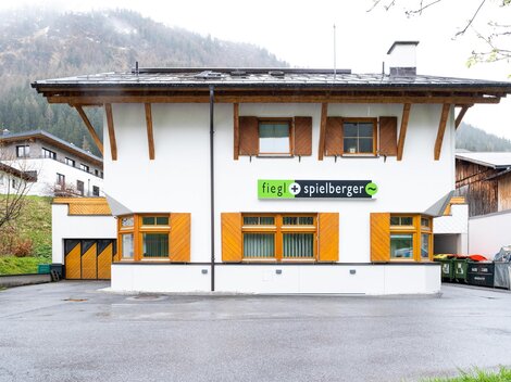 Ein modernes Gebäude mit einem weißen Außenanstrich und Holzverkleidungen. Das Logo von "fiegL spielberger" ist gut sichtbar über dem Eingang angebracht.