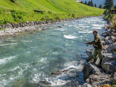 Ein Mann angelt an einem klaren, fließenden Fluss. Umgeben von grünen Wiesen und Bäumen bietet die Landschaft eine ruhige Naturkulisse.