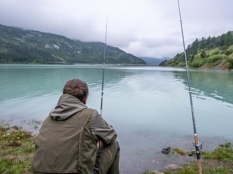 Zwei Angler stehen am Ufer eines klaren Bergsees. Umgeben von grünen Bergen und blauem Himmel genießen sie die Natur.