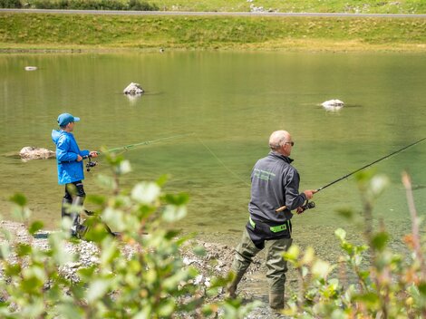 Zwei Angler stehen am Ufer eines klaren Gewässers. Im Hintergrund sind grüne Wiesen und Steine zu sehen.