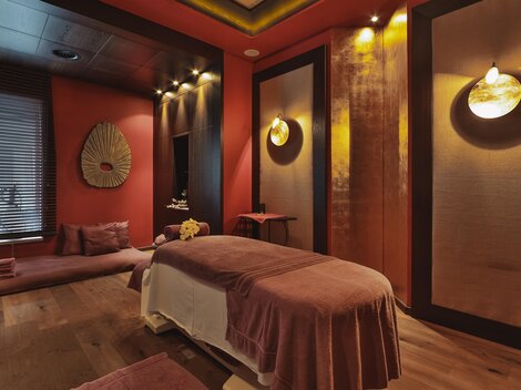 Ein stilvolles Spa-Zimmer mit einer Massageliege und sanfter Beleuchtung. Die Wände sind in warmen Farbtönen gehalten und schaffen eine entspannende Atmosphäre.