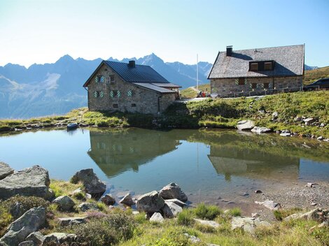 Eine malerische alpine Landschaft mit zwei Hütten am Ufer eines ruhigen Sees. Im Hintergrund erheben sich majestätische Berge unter einem klaren Himmel.