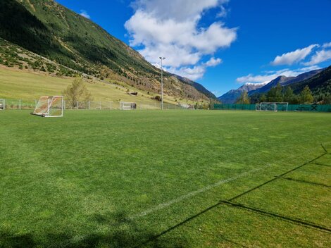 Ein grünes Fußballfeld in einer malerischen Berglandschaft. Im Hintergrund sind hohe Berge und ein klarer blauer Himmel zu sehen.