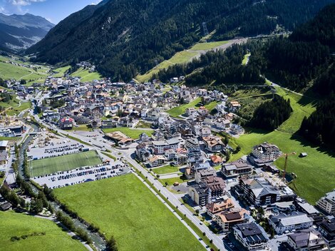 Eine malerische alpine Stadt in einem grünen Tal, umgeben von Bergen. Die Gebäude sind dicht gepackt, und es gibt viele Parkplätze und Straßen.