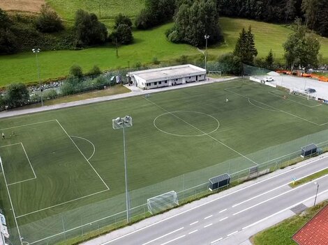 Ein Sportplatz mit Kunstrasen, umgeben von Bäumen. Im Hintergrund sind ein Gebäude und parkende Autos zu sehen.