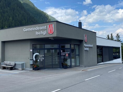 Das Bild zeigt das Gemeindeamt Ischgl, ein modernes Gebäude mit großen Fenstern. Im Hintergrund sind Berge und ein blauer Himmel zu sehen.
