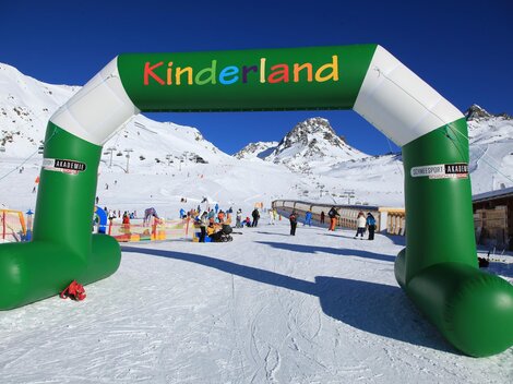 Ein bunter Bogen mit dem Schriftzug "Kinderland" steht in einem verschneiten Skigebiet. Im Hintergrund sind Schlepplifte und Skifahrer zu sehen.