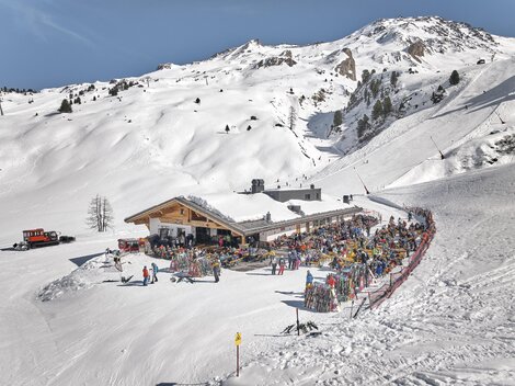 Ein belebtes Skigebiet mit einem großen Restaurant inmitten schneebedeckter Berge. Viele Menschen genießen die Sonne und die Winteraktivitäten.