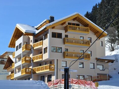 Ein modernes Hotelgebäude mit Holzbalkonen in einer schneebedeckten Umgebung. Im Hintergrund sind Tannenbäume und ein klarer blauer Himmel zu sehen.