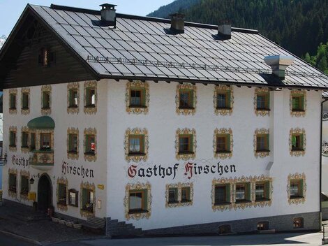 Ein traditionelles Gasthaus mit der Aufschrift „Gasthof Hirschen“ an der Fassade. Das Gebäude hat eine historische Architektur und befindet sich in einer malerischen Berglandschaft.
