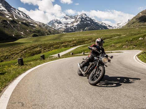 Ein Motorradfahrer fährt auf einer kurvigen Straße in den Bergen. Die Landschaft ist grün mit schneebedeckten Gipfeln im Hintergrund.