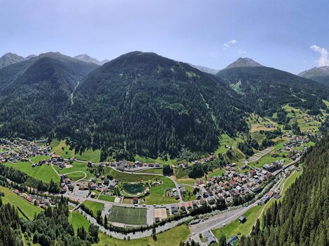 Ein schöner panaromatischer Blick auf ein Tal umgeben von hohen Bergen. Die Landschaft ist grün mit Wäldern und einem kleinen Ort im Hintergrund.
