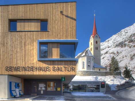 Ein modernes Gemeindehaus mit Holzfassade in Galtür. Im Hintergrund ist eine Kirche mit rotem Kirchturm und schneebedeckte Berge zu sehen.