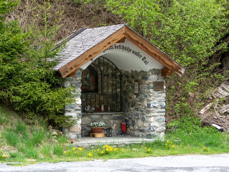Eine kleine Kapelle aus Stein mit einem Holzdach steht in der Natur. Umgeben von Bäumen und Gras bietet sie einen ruhigen Rückzugsort.