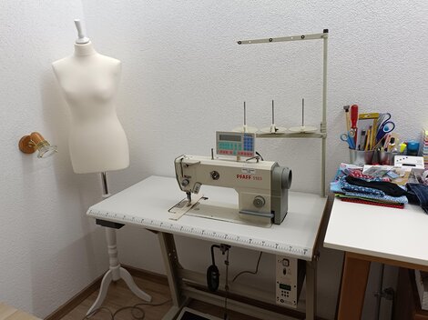 Ein heller Nähraum mit einer Nähmaschine und einer Schneiderpuppe. Auf dem Tisch sind verschiedene Nähutensilien und Stoffe sichtbar.