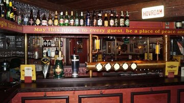 A cozy bar with a long counter and many drinks. Above the counter hang various bottles and a sign with a friendly message.