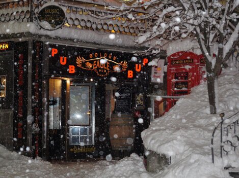 A cozy pub named "Golden Eagle" is hidden in the snow. The snowflakes fall thickly as the warm light of the entrance seems inviting.
