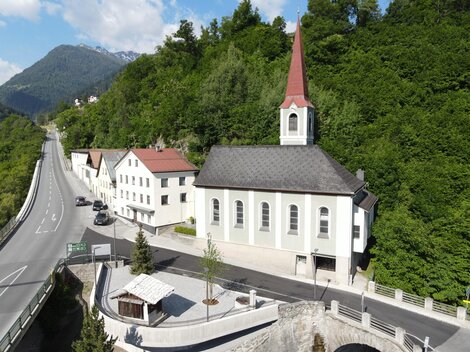 Eine kleine Kirche mit einem roten Dach und einem hohen Turm steht an einer Straße. Umgeben von grünen Hügeln und Wohngebäuden.
