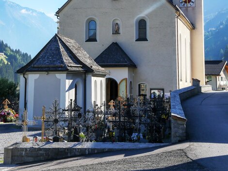 Eine malerische Kirche mit einem charakteristischen Turm in den Alpen. Im Vordergrund befindet sich ein kleiner Friedhof.