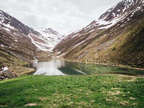 Ein ruhiger Bergsee umgeben von schneebedeckten Bergen. Die Landschaft ist grün und idyllisch.