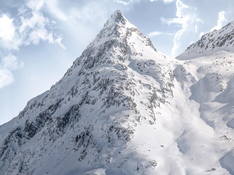 Ein majestätischer Berg mit schneebedeckten Hängen unter einem bewölkten Himmel. Die Gipfel sind in strahlendes Sonnenlicht getaucht.