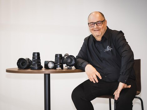Ein lächelnder Mann sitzt an einem Tisch und präsentiert mehrere Kameras und Objektive. Er trägt ein schwarzes Hemd und wirkt freundlich und kompetent. | © Bernhard Gruber – Photography