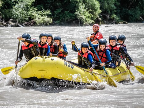 Eine Gruppe von acht Personen paddelt in einem gelben Raft auf einem Fluss. Sie tragen Helme und Schwimmwesten und haben viel Spaß.