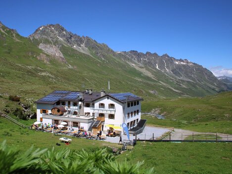 A charming mountain house in a green landscape. In the background, majestic mountains and a blue sky are visible.