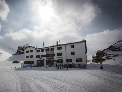 Ein weißes Gebäude im Schnee, umgeben von Bergen und Wolken. Menschen sind in der Nähe des Hauses zu sehen, das eine gemütliche Atmosphäre ausstrahlt.