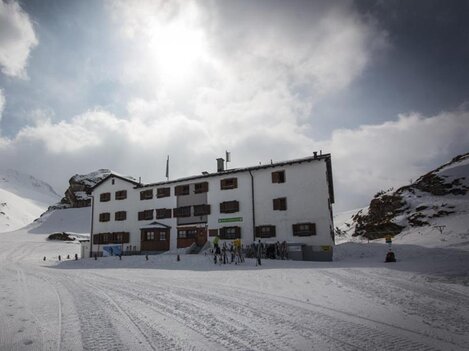 A white building in the snow, surrounded by mountains and clouds. People can be seen near the house, which radiates a cozy atmosphere.