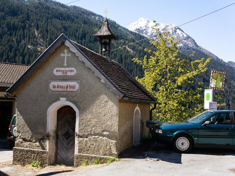 Eine kleine Kapelle mit einer eleganten Dachform und einem Kreuz. Im Hintergrund sind Berge und Bäume zu sehen. | © Bernhard Gruber – Photography