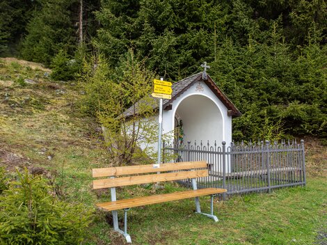 Eine kleine Kapelle steht in der Natur, umgeben von grünen Bäumen. Neben der Kapelle befindet sich eine Holzbank zum Sitzen und Ausruhen. | © Bernhard Gruber – Photography