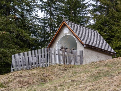 Eine kleine Hütte mit einem Holzzaun in einem Waldgebiet. Der Himmel ist bewölkt, und der Boden ist mit Gras und Laub bedeckt. | © Bernhard Gruber – Photography
